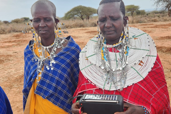 Members of a local community from Tanzania wearing traditional dress are looking at a radio and talking.