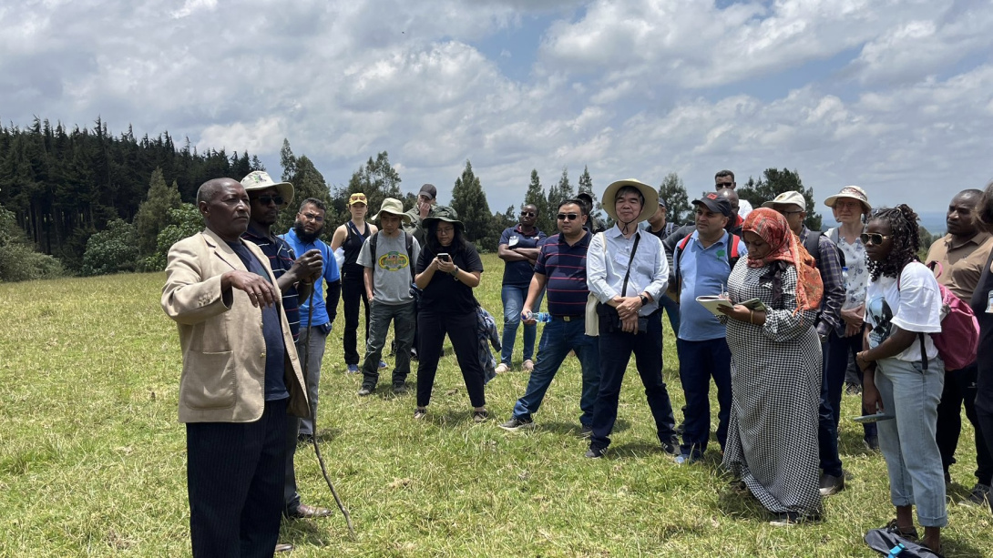A group of people gathered in a green field, listening to their guide.