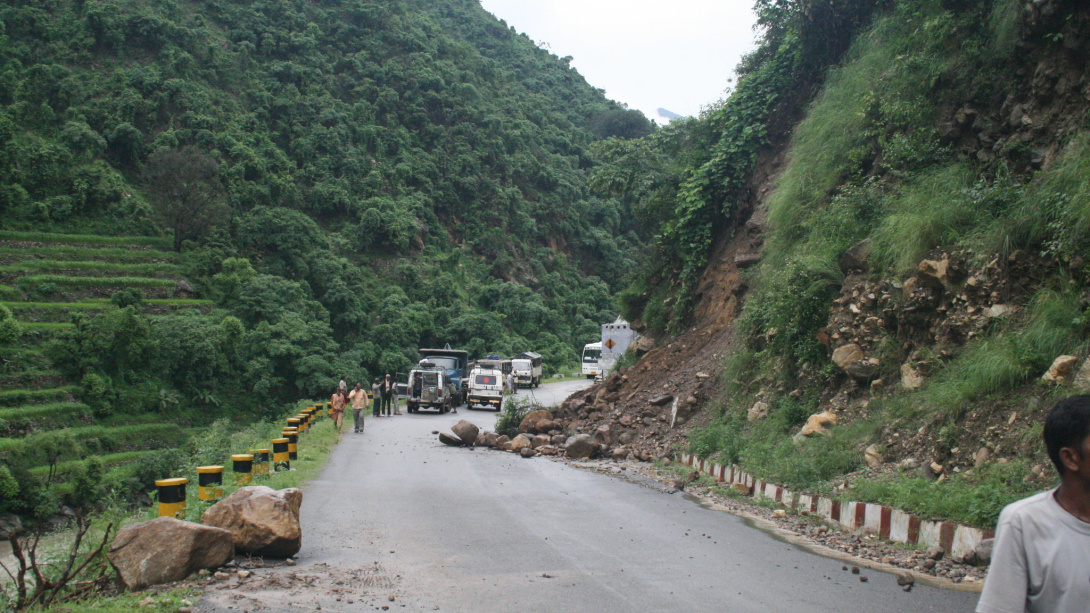 Landslide debris partially blocks a mountain road as vehicles and people gather; lush green hills are visible on both sides.
