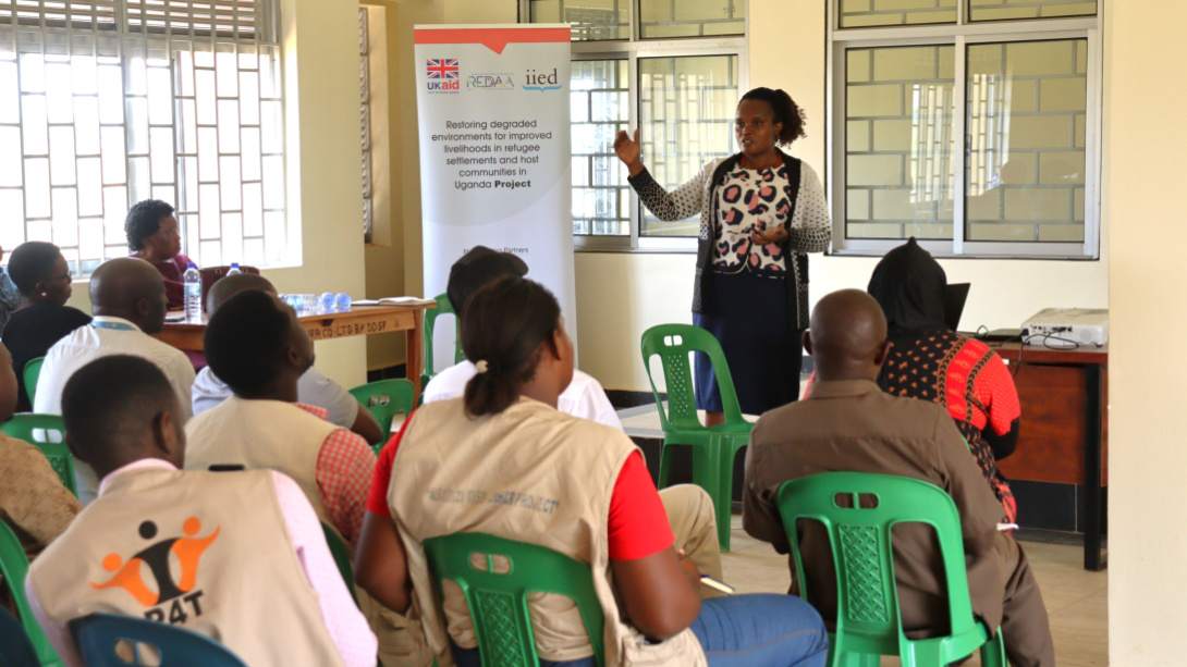 A woman stands in front of a banner and gestures as she speaks to a seated group in a classroom setting. The banner reads 'restoring degraded environments for improved livelihoods in refugee settlements and host communities in Uganda project".